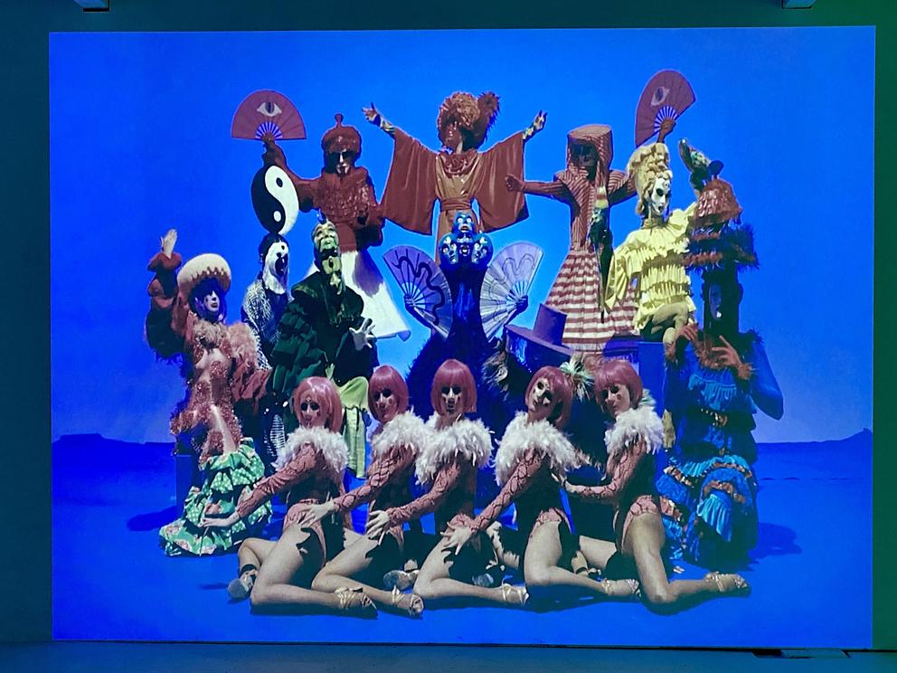 a photo of a film featuring a brightly costume troupe of performers against a blue backdrop