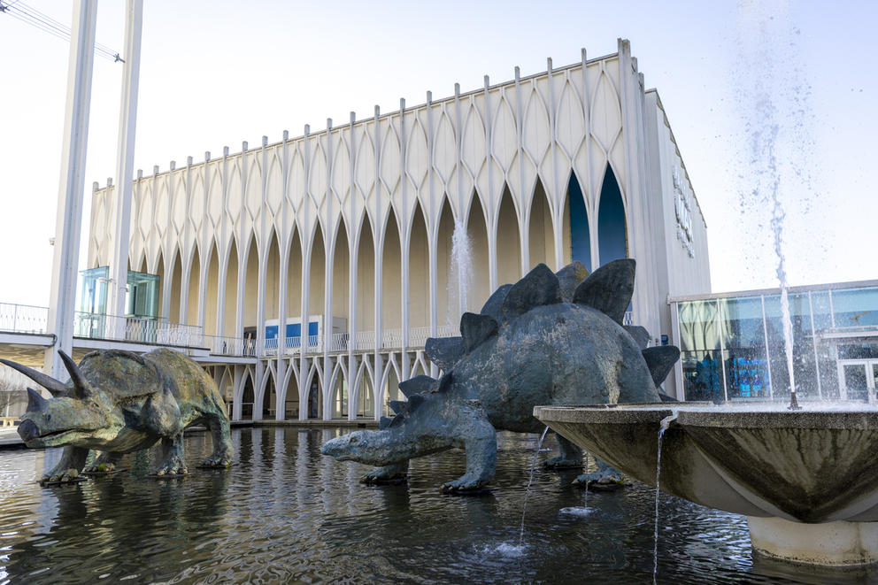 two giant dinosaur sculptures stand in a wading pool with a fountain, in front of a building with a wishbone-like design