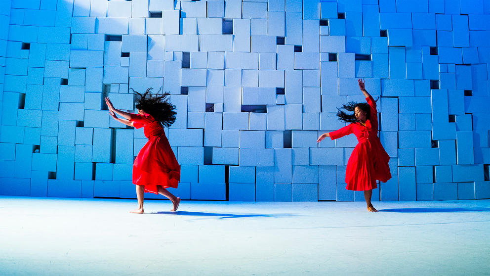 two women in red dresses dancing in front of a white wall piled with boxes