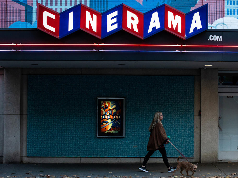 Woman walks her dog in front of a dark facade, up top red and blue squares with white letters spell out CINERAMA