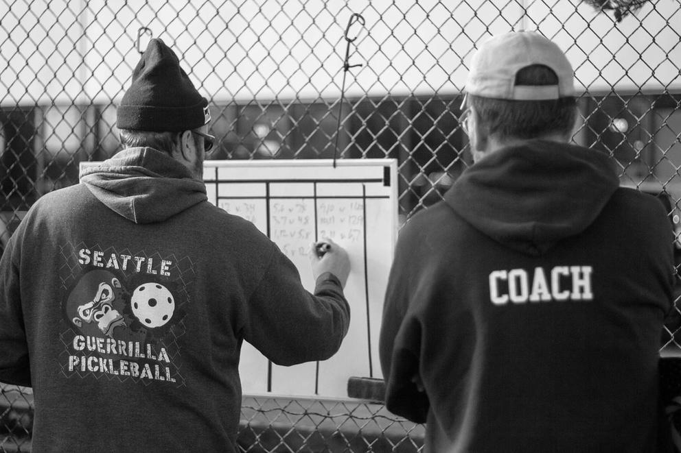 a medium shot of two men with their backs to the camera, one writes on a white board hung on a chainlink fence