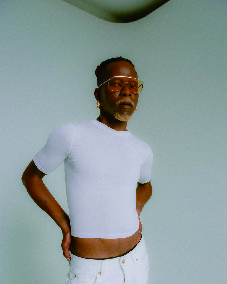 Pierre Kwenders looks at the camera in a white t-shirt, white pants and yellow-tinted sunglasses with a hand on his hip.