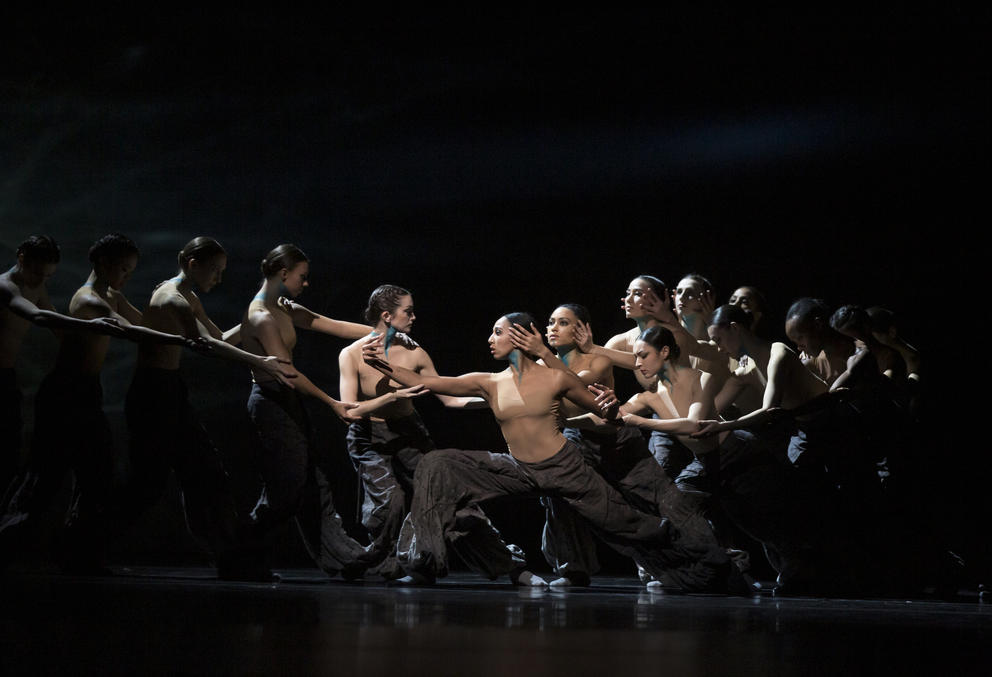 photo of dancers on a dark stage in two lines both facing a central dancer