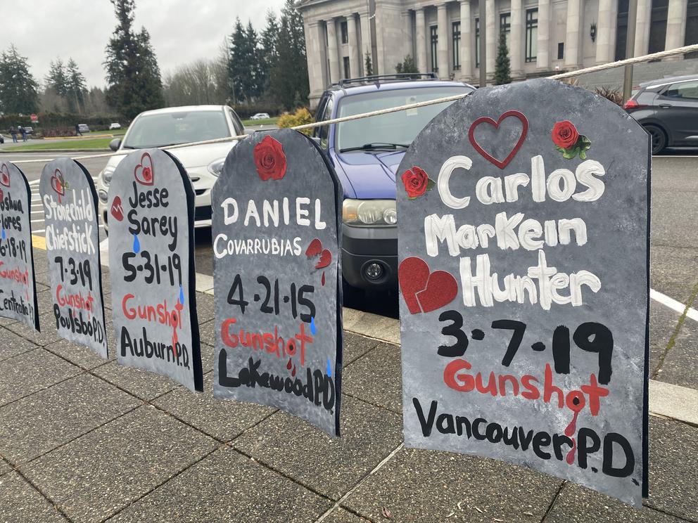 Four signs in the shape of gravestones bear the names of people killed by police and the dates they died. From right to left:  Carlos MarKein Hunter, 3-17-19, Gunshot, Vancouver PD; Daniel Corrubias, 4-21-15, Gunshot, Lakewood PD; Jesse Sarey, 5-31-19, Gunshot, Auburn PD; Stonechild Chiefstick, 7-3-19, Gunshot, Poulsbo PD.