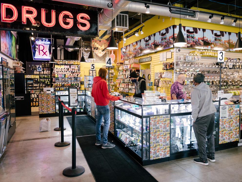 Customers approach a display case filled with marijuana products. At left in the background a sign reads "drugs" in neon letters