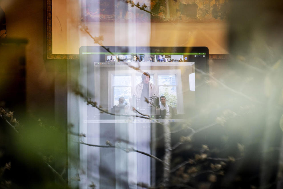 Ellis Gould is seen on a computer screen through a window reflecting trees outside as he conducts his virtual bar mitzvah