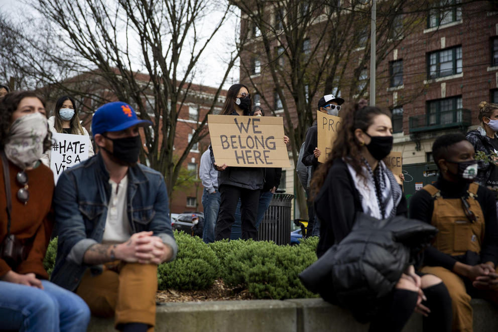 masked people gather at an outdoor rally, a central figure holds a sign that reads "We Belong Here"
