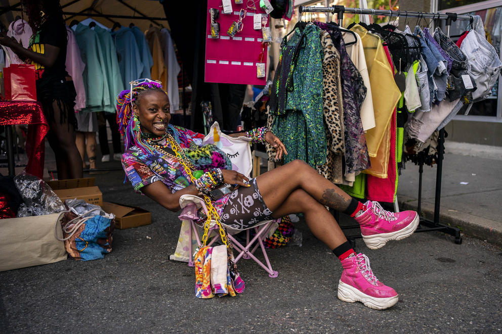 Andreya Taylor reclines in a camp chair wearing pink shoes colorful clothing. A rack of more colorful is behind her. 