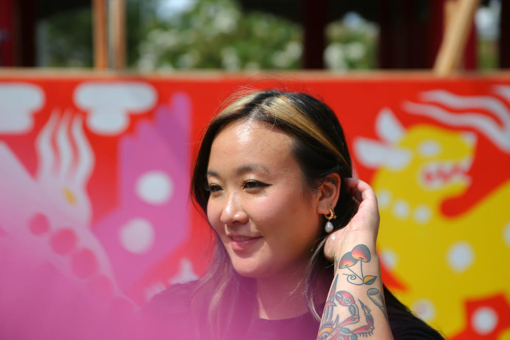 Stevie Shao smiles and tucks her hair behind her ear while standing in front of a colorful mural