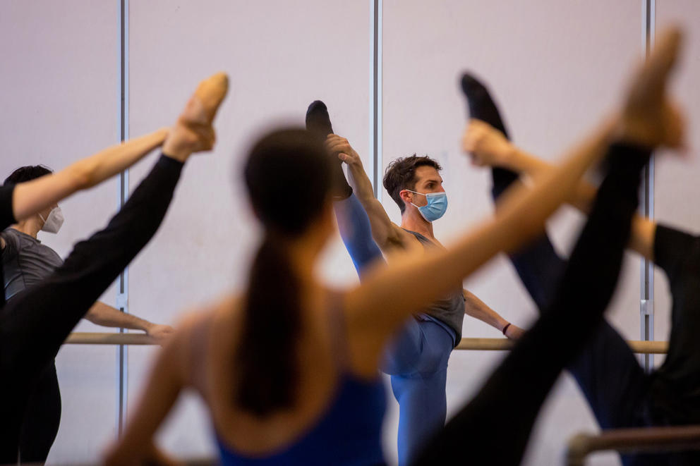 A masked Lucien Postlewaite is seen through the raised legs and arms of other Pacific Northwest Ballet dancers as they warm up at the barre