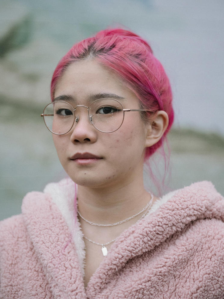 A close up portrait of Alex Su, who stares intently at the camera. She has pink hair, a pink sweater and glasses speckled with rain drops.