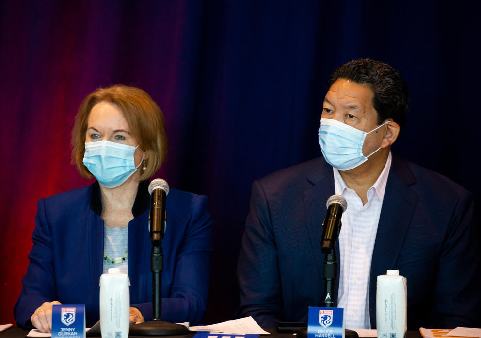 Jenny Durkan and Bruce Harrell sit side by side in front of a neutral backdrop wearing identical blue surgical masks at a press conference