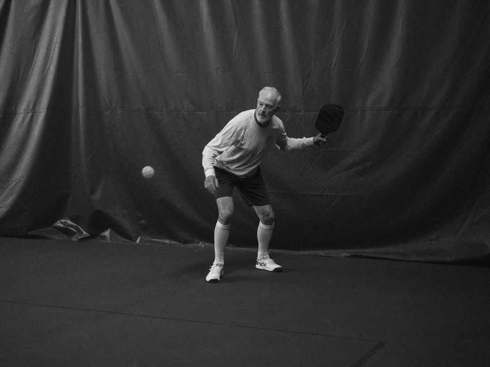 A black and white photo of a man with white hair playing pickleball, the ball is midair and a curtain drapes behind him