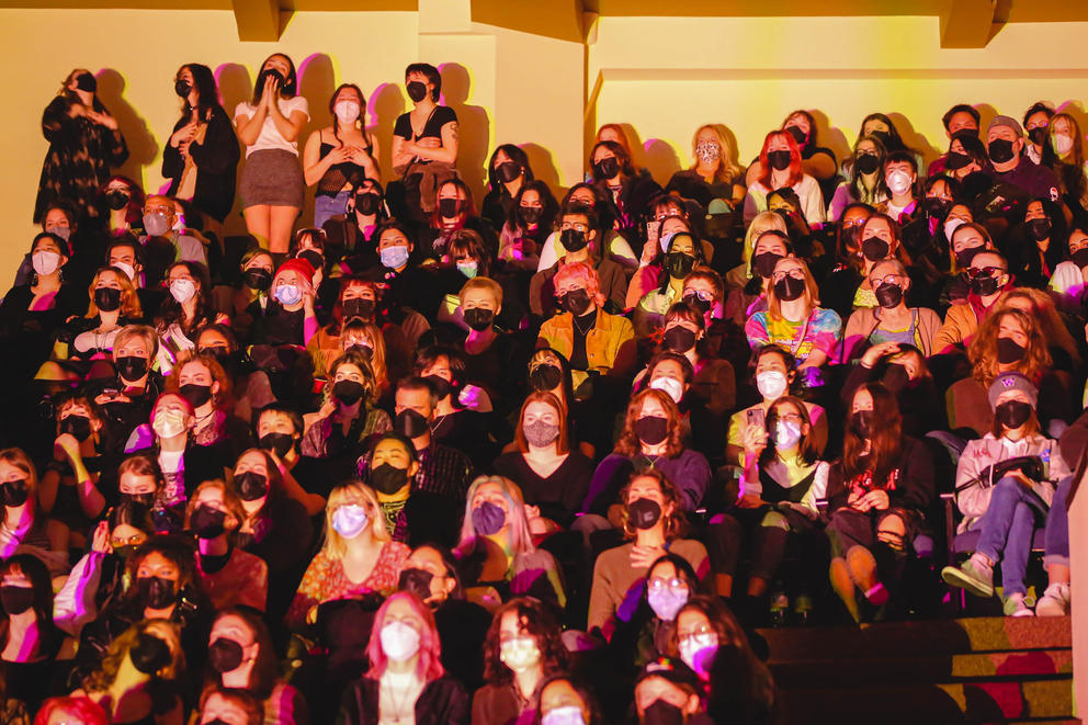 An audience wears masks and is lit up by colored stage light