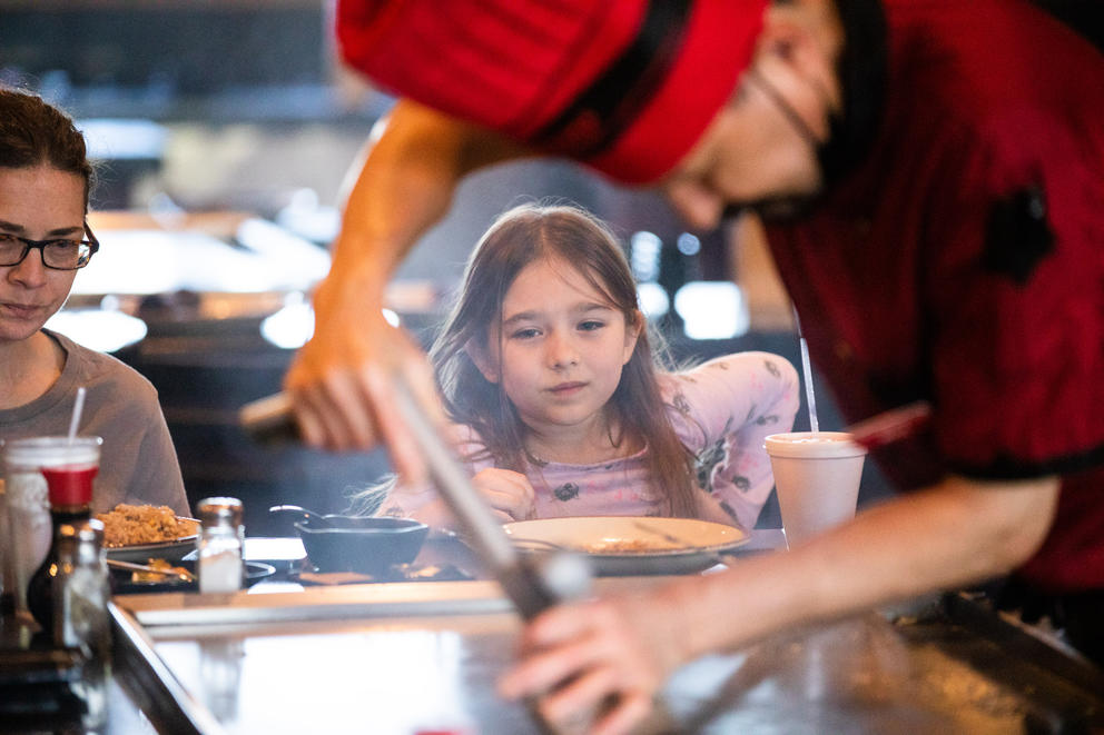 A young girl is seen through the arms of a chef in a red chef's hat as he makes food