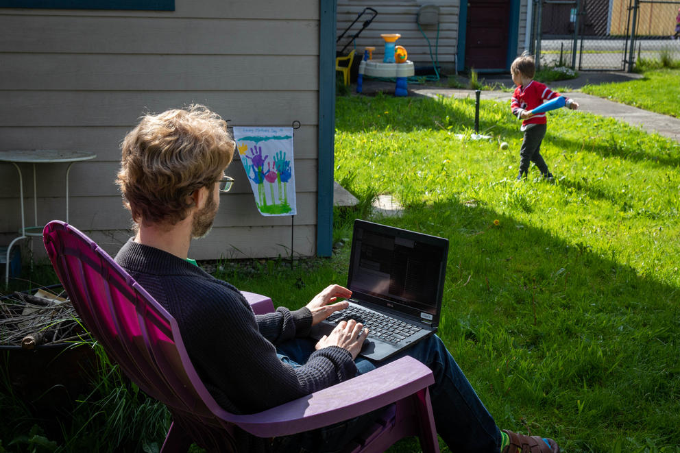 A man sits outside in a deck chair with a computer on his lap, in the background his son plays with a plastic t-ball bat