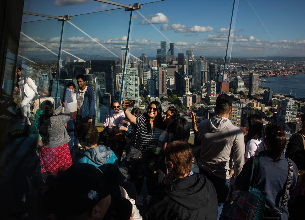 A crowd of people is gathered on the viewing deck of the Space Needle, the Seattle skyline is seen in the background. People are taking selfies. 