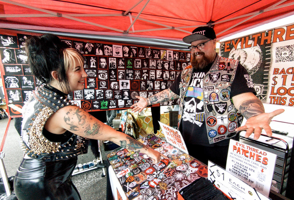 Two people with tattoos and studded and patched vests great eachother in a booth full of patches