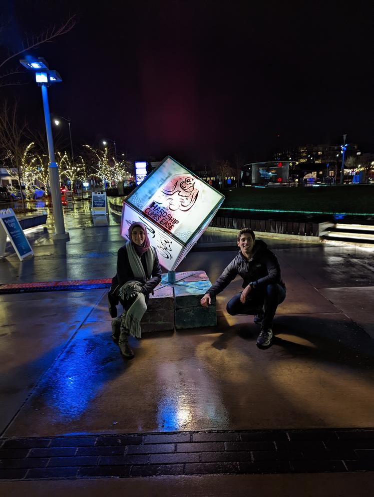 two people pose with a cube that glows with lights from within, t hey sit crouched on either side. in the background is a city scape lighting up the dark night sky