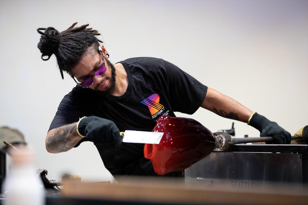 A person with a black t-shirt and dreadlocks, wearing protective glasses, is working on some hot glass