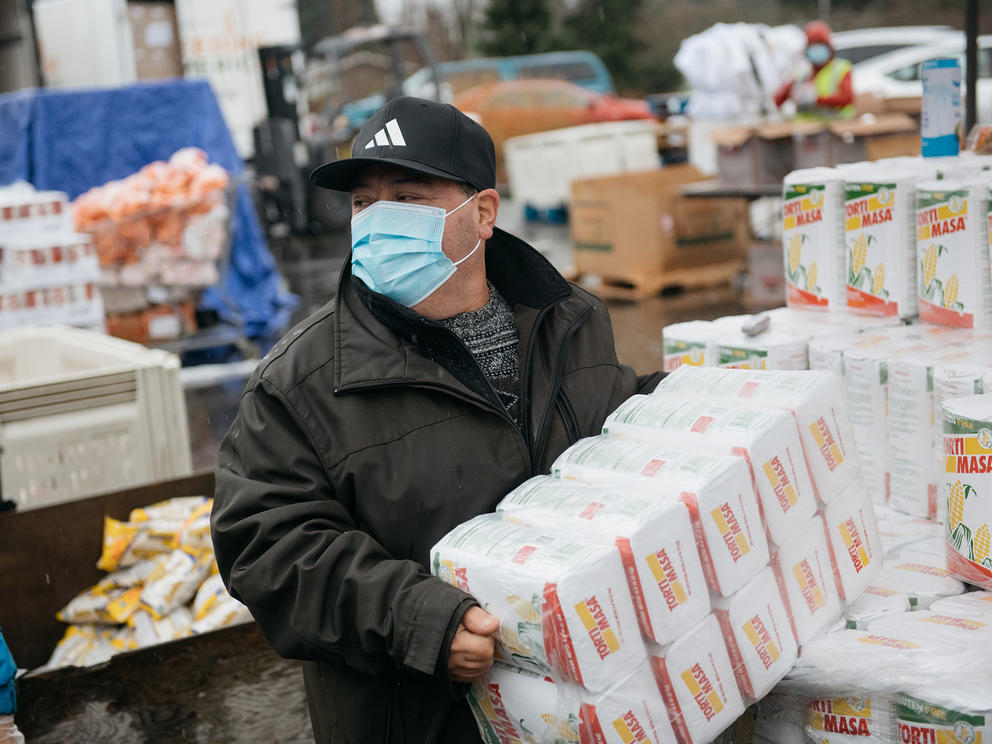 A food bank volunteer carries an armload of masa while wearing a surgical mask.