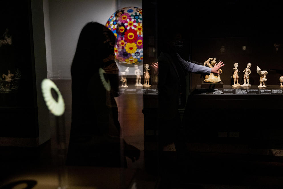 darkened gallery, left: colorful contemporary work, a large disk with cartoon-like figures, right: a variety of small stone statues