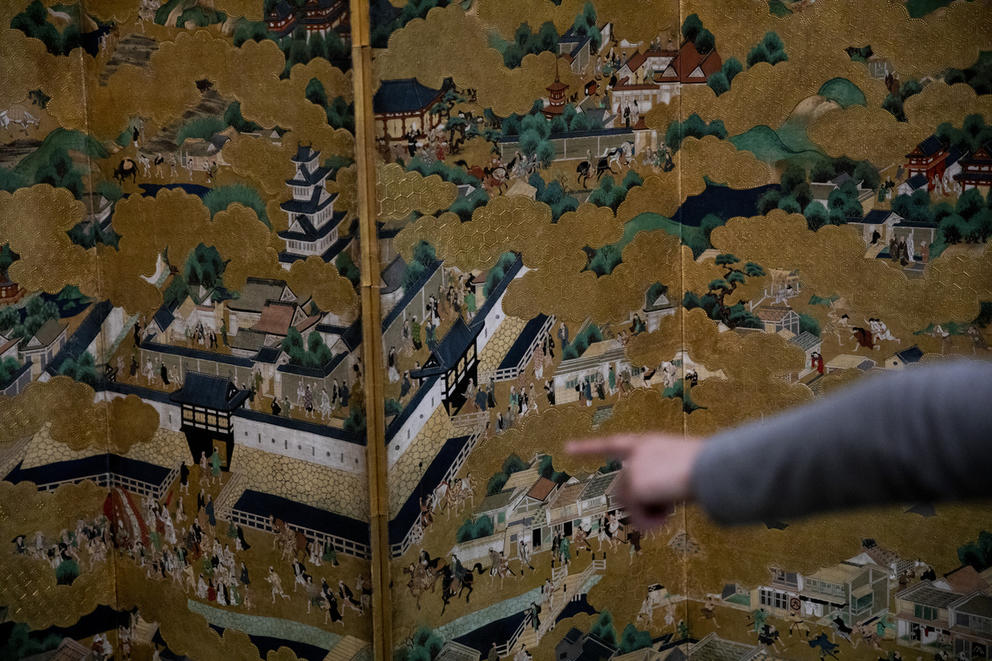hand pointing to a painted panel filled with golden clouds and small figures