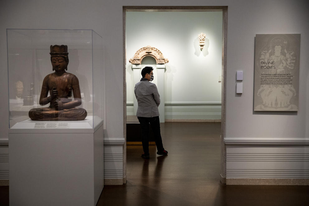 Person walks in museum, left: buddha statue in a glass case on a podium