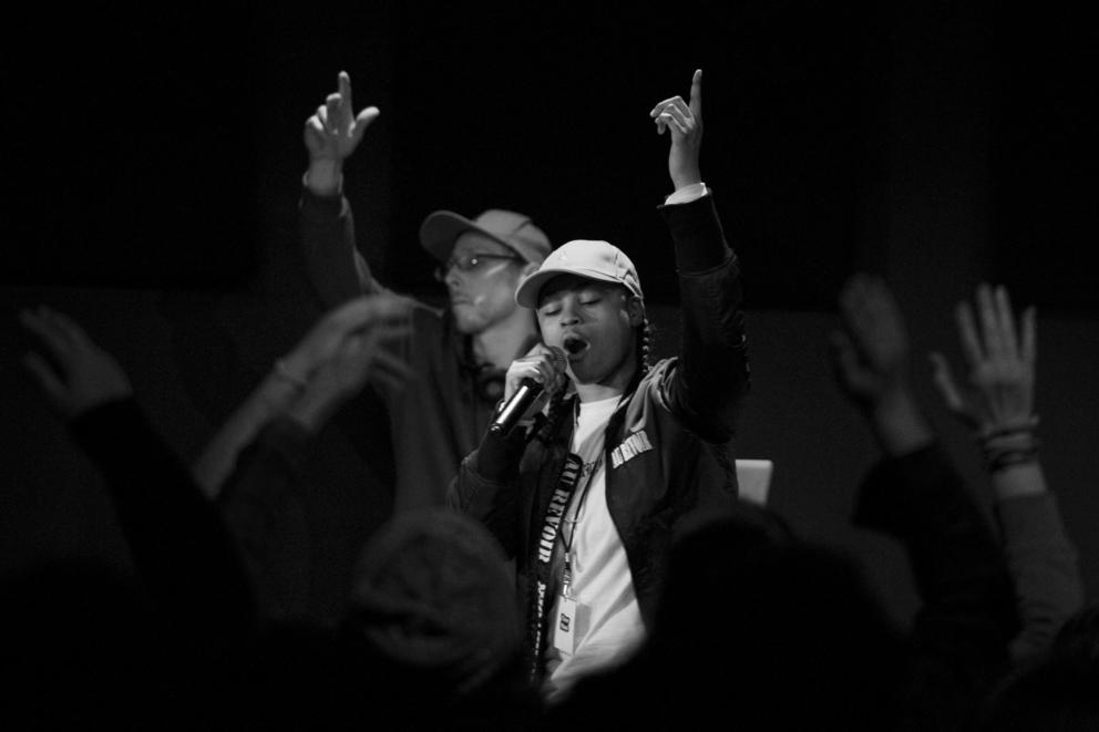 Black-and-white photo of a young person wearing a hat and a white t-shirt with their hand up in the air and a microphone in front of their mouth