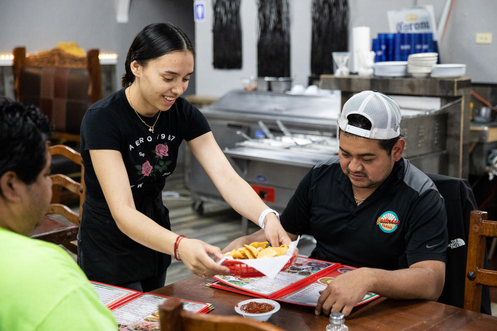 A woman serves Mexican food to two people sitting at a table. 
