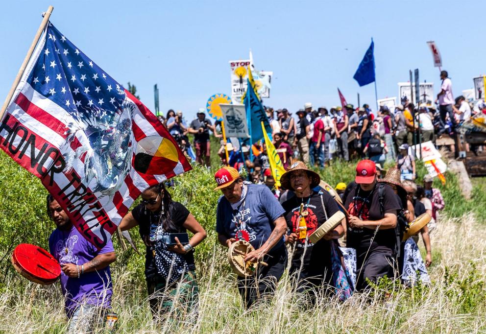 A group of Indigenous activists walk through tall grass holding flags and signs, one of which reads "honor treaties" on a painted american flag