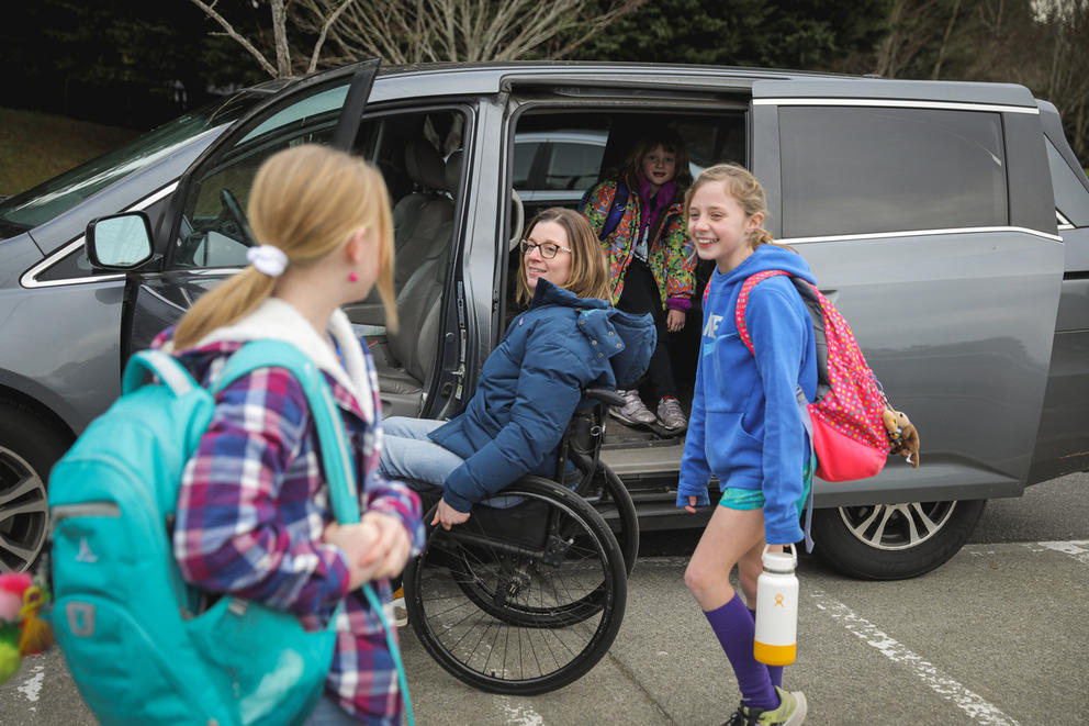 Two children get into a van and wave goodbye to a friend, as their mother rolls up to the driver's seat in her wheelchair.