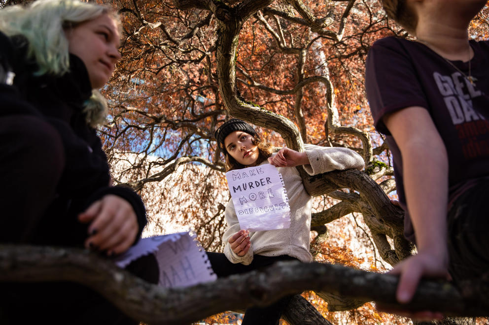 A student leaning against a tree holds a sign saying "Make Murder More Difficult."