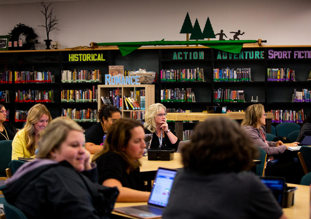About eight female school employees sit at tables near rows of bookshelves listening to a presentation.