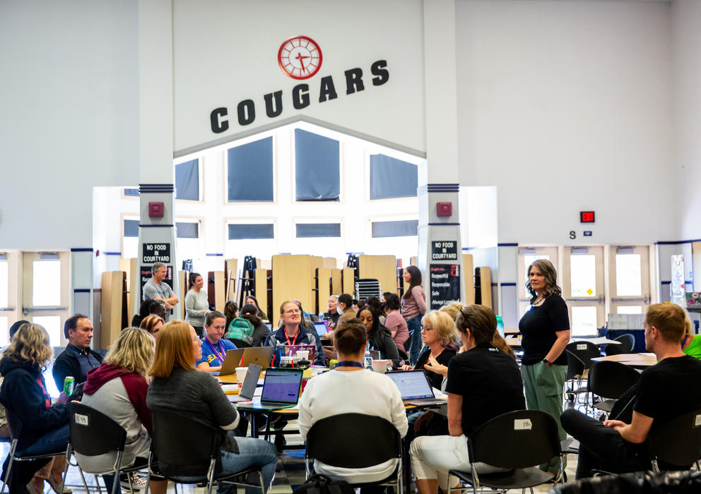 About a dozen school employees sit around a circular table with laptops as the word "Cougars" can be read above their heads on the far wall. 