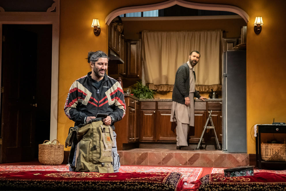 a stage play setting with one man packing a backpack, the other downstage in a kitchen