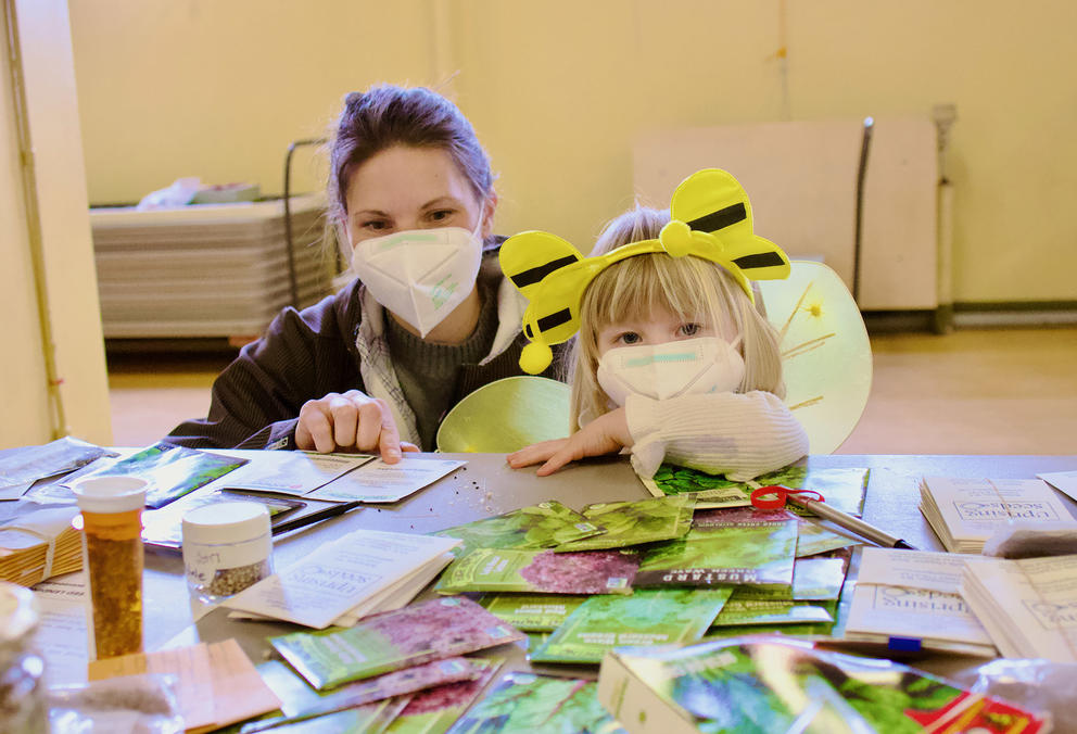 A woman and her daughter who is wearing a bee costume, are both masked and crouched behind a table with seed packets on it