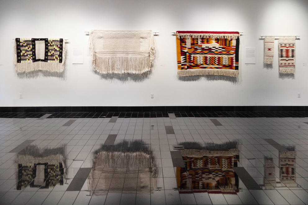 Weavings on display on a white gallery wall, reflected in the shiny black floor below