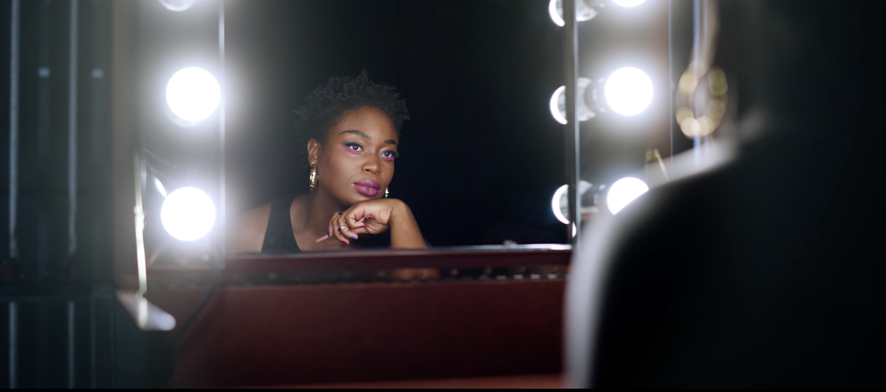 a black woman with dramatic eye shadow looks at herself in a backstage theater mirror