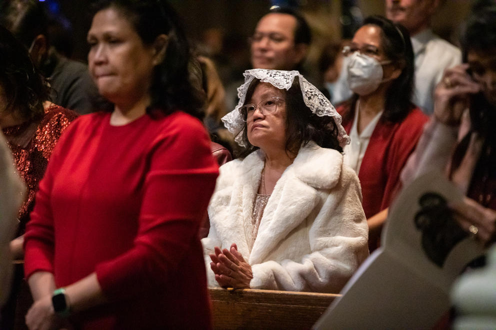 A woman in white folds her hands in a pew surrounded by other parishoners