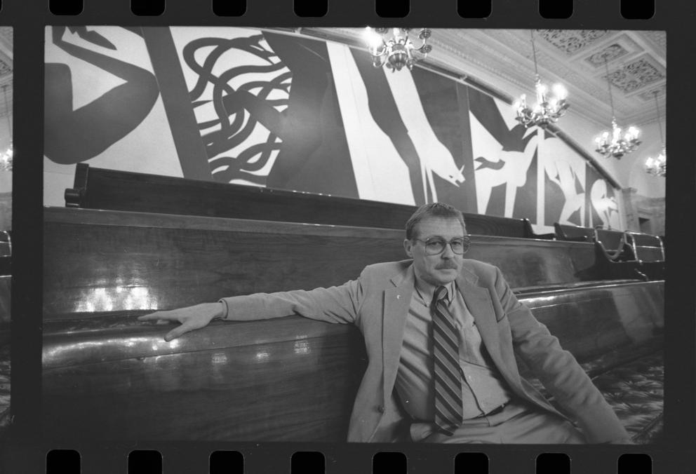 a black and white photo of a man in a suit and glasses, sitting in front of large geometric murals