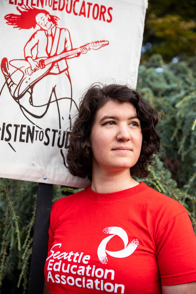 A teacher wears a red T-shirt that says "Seattle Education Association," in front of a white sign that says "Listen to Educators, Listen to Students.""