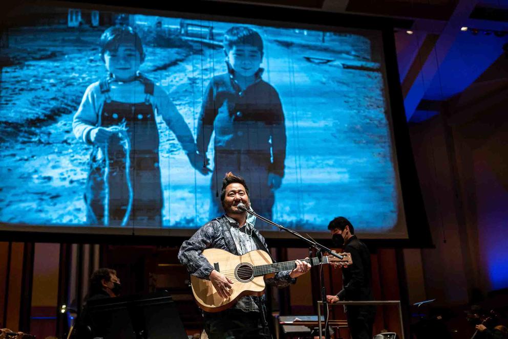 photo of a man holding a guitar and smiling in front of a large screen showing a vintage photo of two young children
