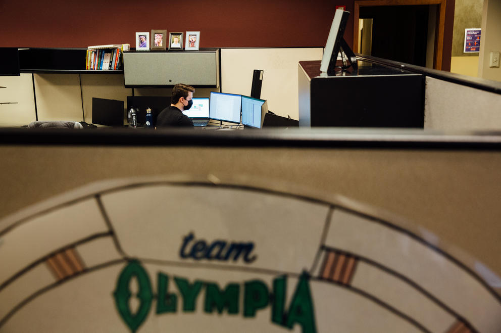 Dylan Wharton, an assistant auditor, faces away as he works at his computer in his cubicle. A sign in the foreground reads "Team Olympia."