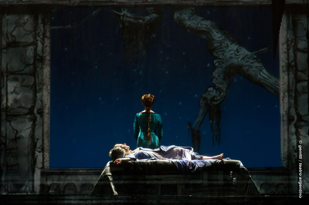Dark stage with person lying down and one person with back turned in blue dress