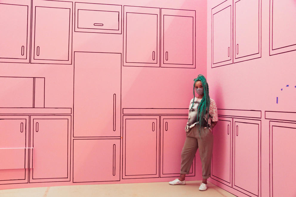 woman with blue hair standing in between pink walls painted as a kitchen wall