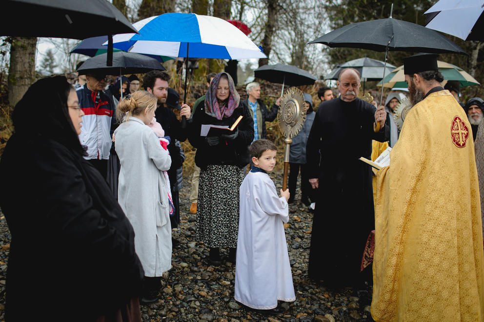 A group of people stand outside under umbrellas. An altar boy in white is at the center of the crowd.