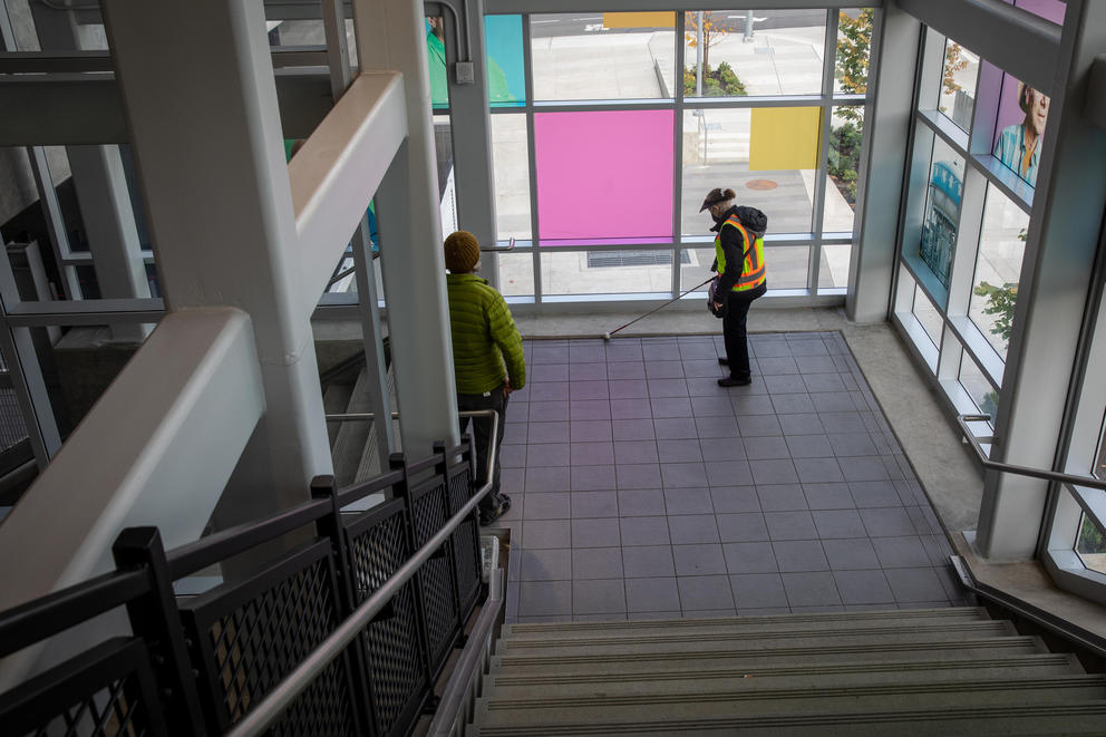 A very wide shot of Lynn Chase, wearing a neon safety vest and black visor, working her way down a staircase using her cane at the Northgate light rail station. Orientation and mobility trainer David Miller wearing a green jacket watches from just a few feet away.