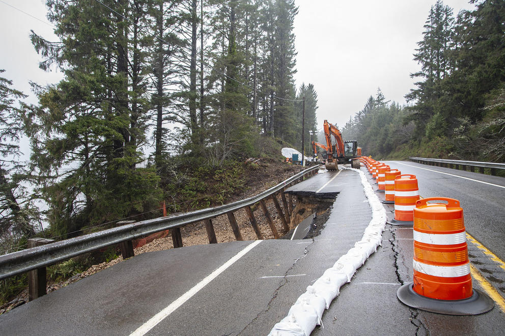 Large orange pilons line a highway where a chunk of the road has fallen down an eroding hillside