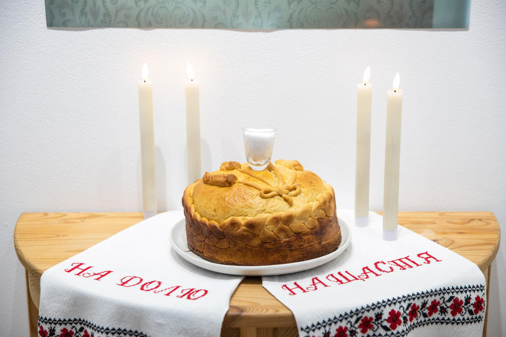 A round loaf of bread sits on a piece of fabric on a table, flanked by two candles on each side. On the fabric, red letters spell out: “na dolju” and “na shastja." 